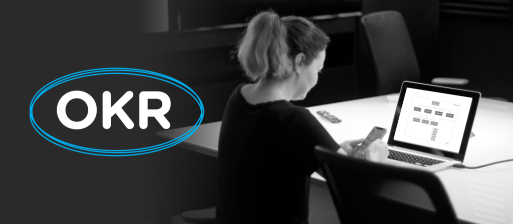 Uma mulher de costas usando camiseta preta está sentada em frente a uma mesa com um notebook, segurando um celular. Ao seu lado, está a sigla OKR, que foi inserida digitalmente na imagem, circulada por uma forma oval azul.
