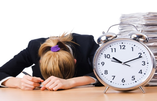 A imagem mostra uma mulher de cabelos loiros com o rosto sobre uma mesa de trabalho e ao lado dela há um relógio analógico