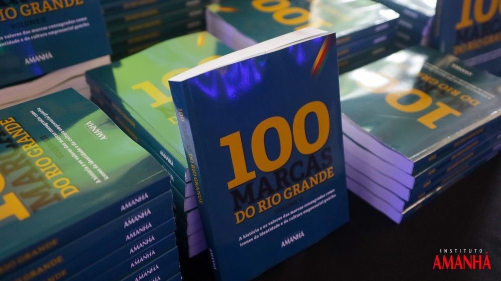 Exemplares do livro "100 Marcas do Rio Grande - Volume II" empilhados em cima de uma mesa, com um deles de pé e a capa voltada para frente.