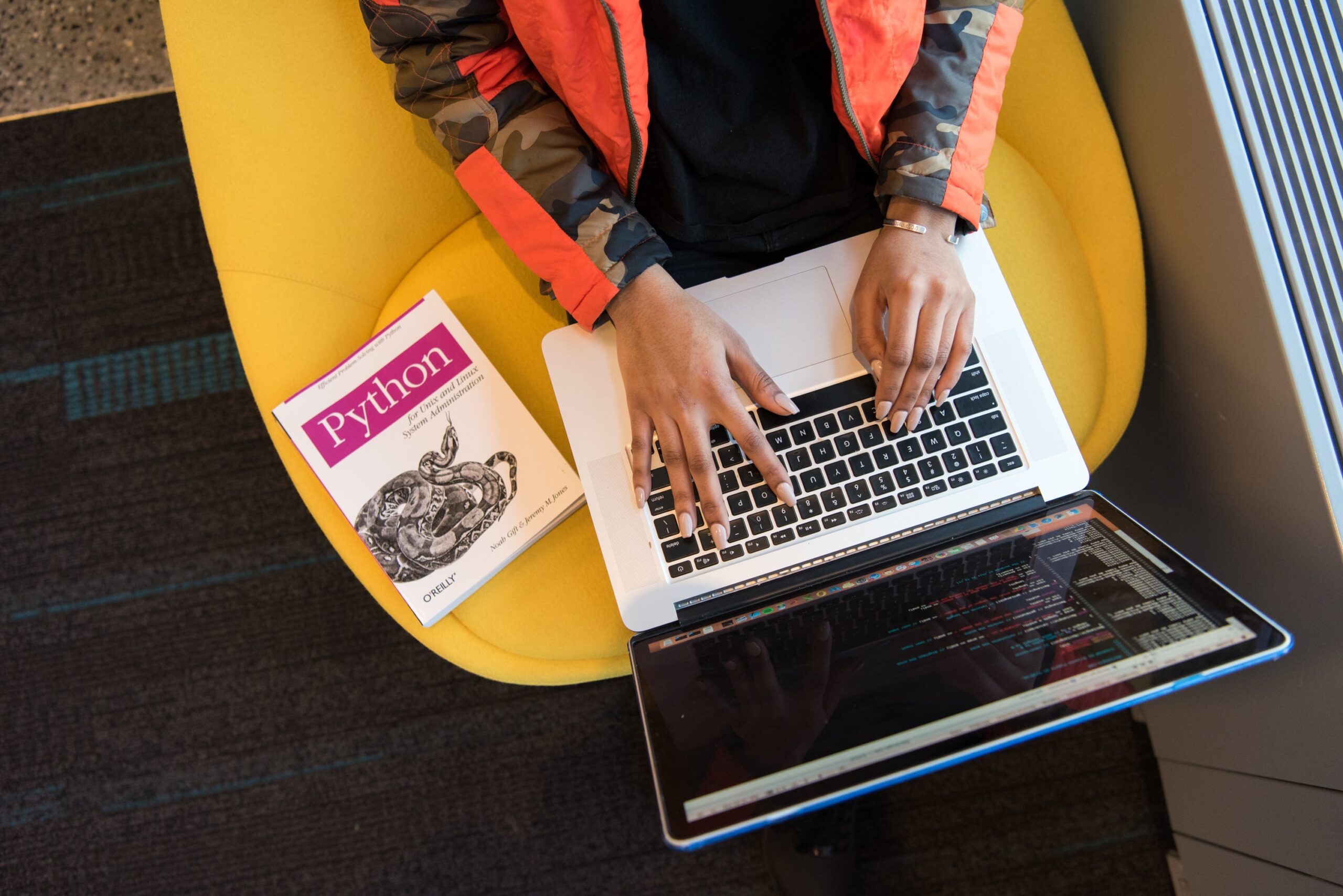 Mulher de pele negra escrevendo código em um MacBook. Ela está sentada em uma poltrona de cor amarela, veste uma jaqueta de cor laranja e tem um livro de Python ao seu lado.