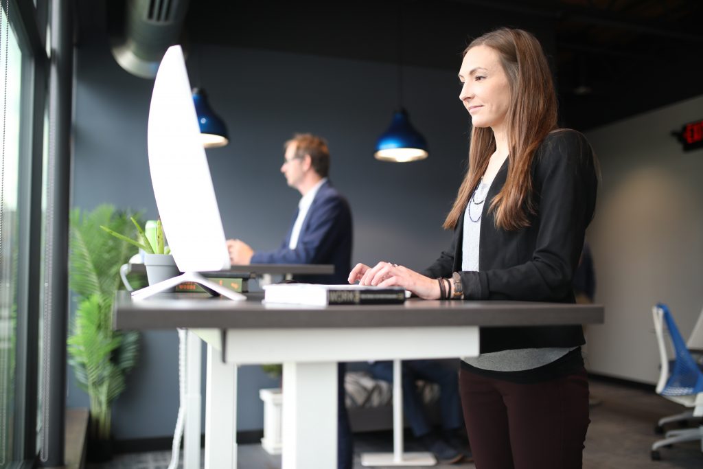 A imagem mostra uma mesa de escritório com dois computadores e na frente dela há duas pessoas em pé.