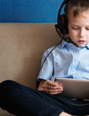 Um menino em um sofá olha para um tablet e usa fones de ouvido.