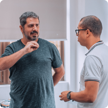 Two men talk in an environment with white walls, with a window in the background.