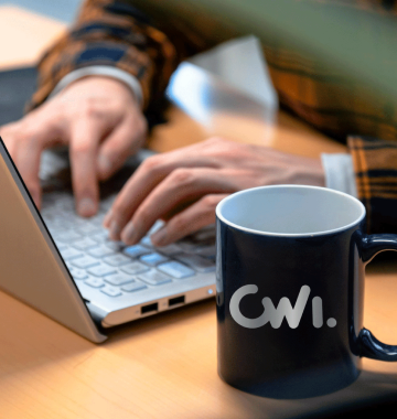 Ao lado de mãos trabalhando em um notebook, uma caneca preta com o logotipo da CWI prateado.