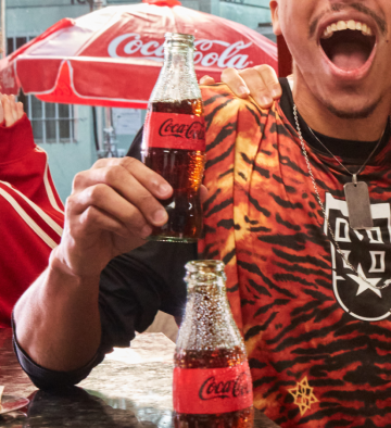 Homem sorridente segura uma garrafa de Coca-Cola em ambiente descontraído com guarda-sol da marca ao fundo.