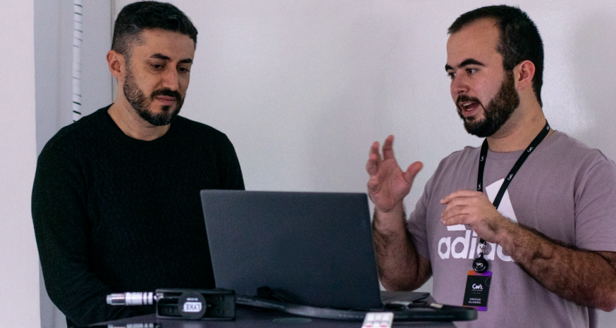Dois homens conversam com um notebook aberto em frente a eles.