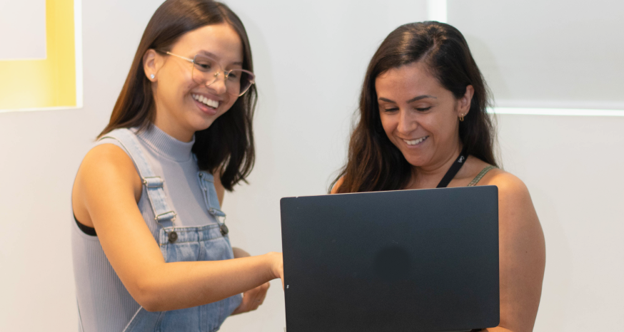 Duas mulheres conversam em frente a um notebook aberto.