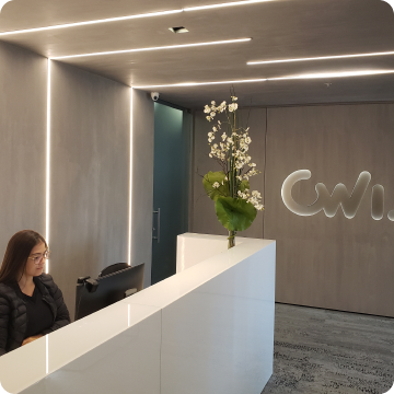 A imagem mostra uma mulher branca sentada em frente a um computador. Ao fundo há o logo da CWI.