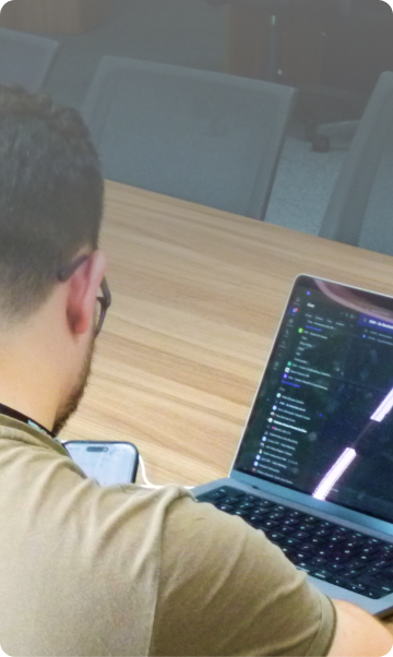 Homem de costas com smartphone na mão em frente a um notebook aberto.