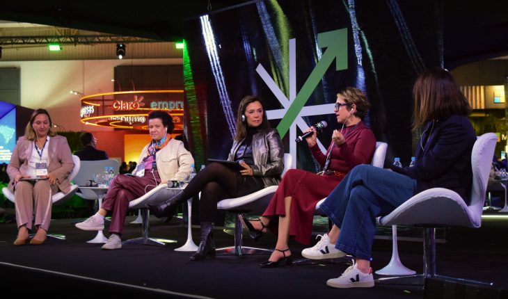 A imagem mostra um paco do evento Febraban Tech. Nele, há cinco mulheres sentadas em cadeiras. Uma delas está falando em um microfone enquanto gesticula com a mão.