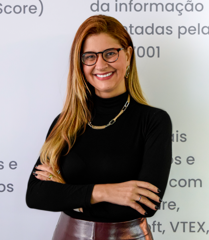 Uma mulher de cabelo louro, óculos de aro preto, blusa preta de gola alta, saia vermelha está com os braços cruzados e sorrindo para a foto.