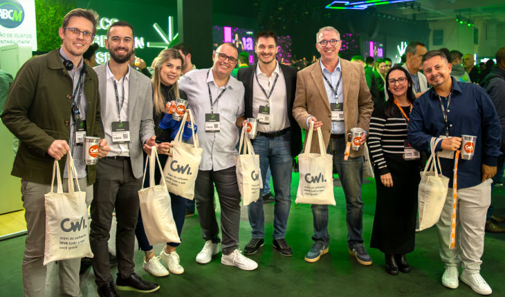 Na imagem, um grupo de pessoas posa para a foto sorrindo e segurando brindes da CWI. O fundo mostra o show de happy hour do Febraban Tech.