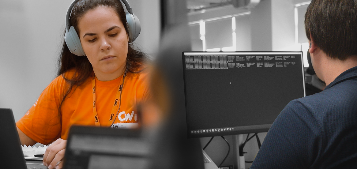 Pessoa com fones trabalhando no notebook enquanto outra programa em tela preta ao fundo.