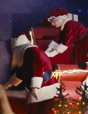 Personagens vestidos de Papai Noel organizam presentes em um trenó iluminado, em uma cena natalina com clima festivo.