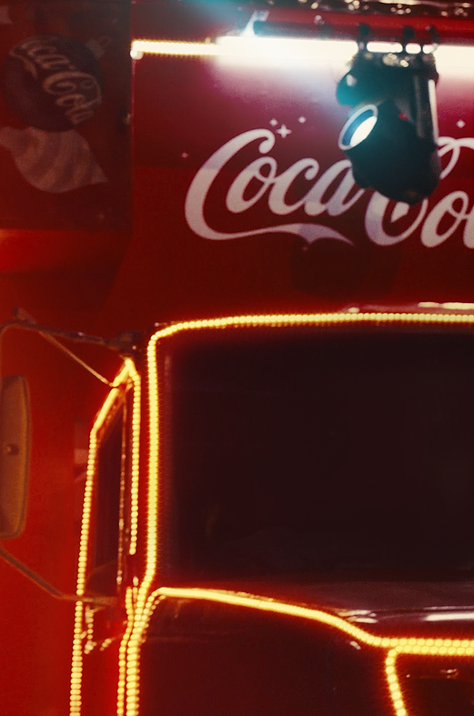Imagem em close de uma estrutura vermelha com a marca Coca-Cola em destaque, contornada por luzes de LED laranja. Há iluminação cênica no topo, sugerindo um ambiente noturno ou de evento promocional.