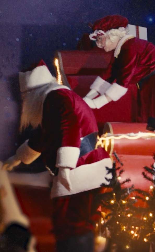 Personagens vestidos de Papai Noel organizam presentes em um trenó iluminado, em uma cena natalina com clima festivo.