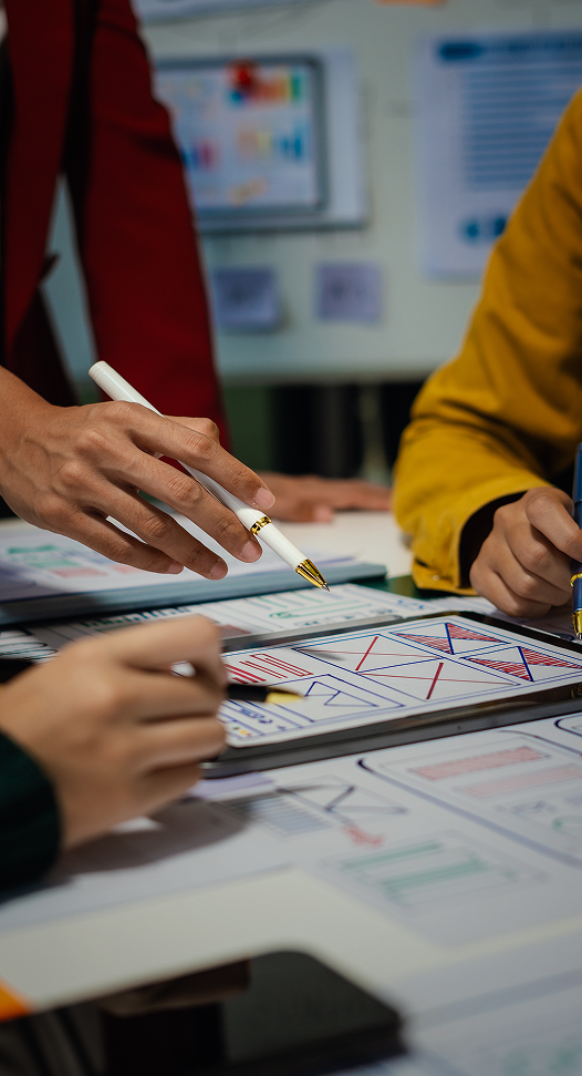 Sobre uma mesa estão papéis e um tablet e, acima disso, aparecem mão de pessoas segurando canetas. Ao fundo, estão boards comumente usados em organização de projetos.