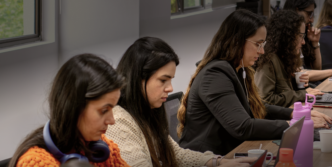 cwisers aparecem sentados(as) lado a lado em uma mesa, todas as pessoas estão com a cabeça baixa olhando para seus notebooks enquanto trabalham.