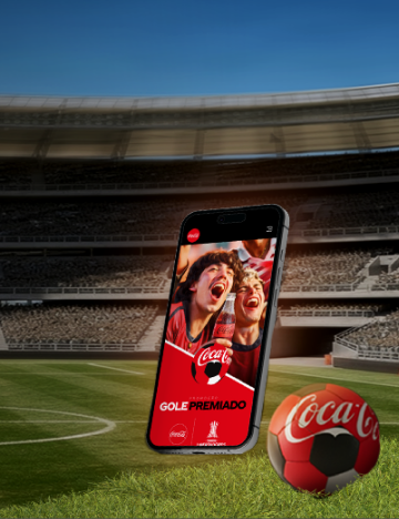 O fundo mostra um estádio de futebol e, sobre o gramado, está um celular e uma bola de futebol da Coca-Cola.