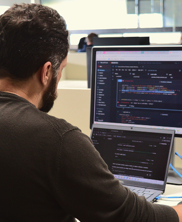 Sobre um fundo na cor clara, há a imagem de um homem visto de costas, sentado em uma mesa de trabalho, usando um notebook conectado a um monitor externo. Nas telas, há códigos de programação em fundo escuro.
