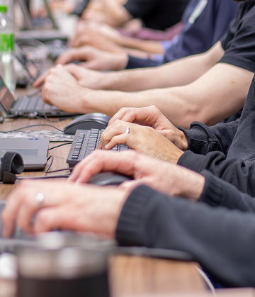 Sobre um fundo na cor clara, há a imagem de dois homens sentados lado a lado em uma mesa de trabalho, usando headsets com microfone e trabalhando em notebooks. Eles estão concentrados, olhando para as telas.