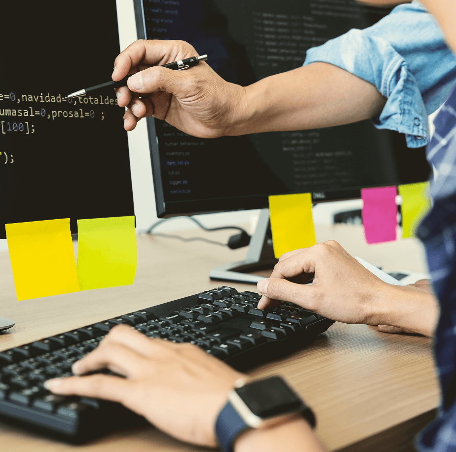 Profissional aponta para código na tela, com post-its coloridos no monitor, em ambiente de desenvolvimento.