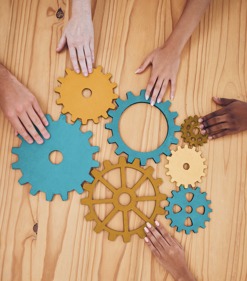 Vistas de cima, diversas mãos de tons de pele variados estão posicionadas sobre uma mesa de madeira clara, cada uma tocando ou movendo engrenagens coloridas que se conectam entre si. As engrenagens possuem diferentes tamanhos e cores, incluindo tons de azul-turquesa, amarelo e dourado, espalhadas pelo centro da composição. A imagem simboliza conceitos como trabalho em equipe, colaboração, processos e o funcionamento conjunto de diferentes partes para um objetivo comum.
