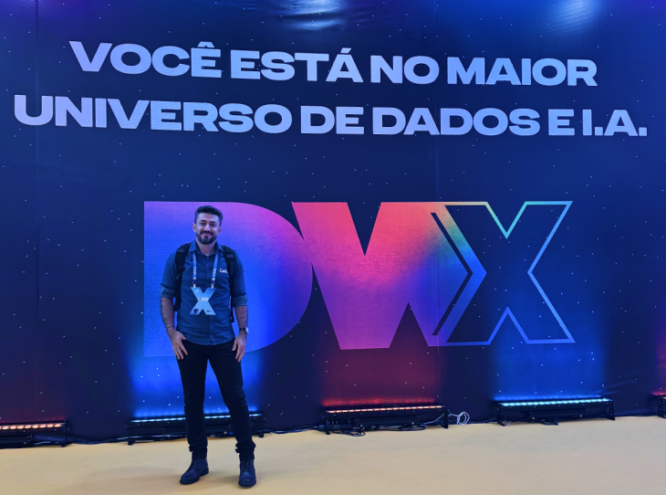 A imagem horizontal mostra André Nunes em pé diante de um grande painel azul escuro com temática espacial e pontos brilhantes que lembram estrelas. André é um homem de pele clara, com cabelos pretos curtos e barba aparada, vestindo uma camisa social azul de mangas dobradas, calça preta e sapatos pretos. Ele usa um crachá pendurado no pescoço e uma mochila preta nas costas. No painel, na parte superior, lê-se em letras garrafais brancas: "VOCÊ ESTÁ NO MAIOR UNIVERSO DE DADOS E I.A.". Logo abaixo, ao centro, destaca-se a sigla "DWX" em letras grandes e estilizadas, com um degradê de cores que vai do azul e roxo ao rosa e laranja. O chão é de uma cor amarela clara, e na base do painel há luzes de LED que iluminam a estrutura de baixo para cima com tons de azul e laranja.