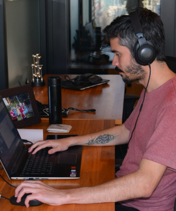Na imagem, um homem de perfil com barba e cabelo escuro aparece sentado em um escritório, focado no trabalho diante de um notebook em uma mesa de madeira. Ele está usando fones de ouvido pretos grandes e uma camiseta de tom avermelhado, revelando uma tatuagem no braço esquerdo enquanto digita no teclado. Ao fundo, sobre a mesa, destacam-se uma garrafa térmica preta, um porta-retratos com uma foto de família e outros objetos de escritório espalhados de forma organizada.