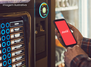 Na imagem, uma pessoa segura um smartphone que exibe o logotipo da Coca-Cola em uma tela inteiramente vermelha, aproximando o aparelho de uma máquina de autoatendimento ou de venda automática. A máquina possui uma tela iluminada com várias opções de produtos e botões azuis na lateral esquerda, além de um sensor circular verde no topo, sugerindo um processo de pagamento digital ou resgate de bebida por aproximação.