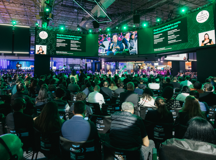 A imagem mostra o evento Febraban Tech: telões espalhados na parte superior com informações sobre a palestra que aparece no palco centralizado. Ao redor, várias pessoas estão sentadas.